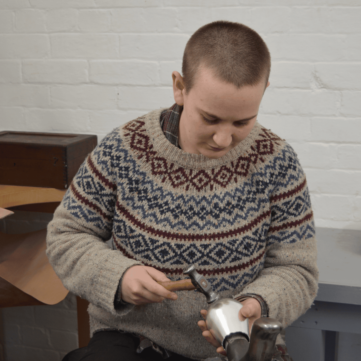Silversmith Annie Higgins, who is taking part in Shine 2026, sitting in front of a white brick wall and using a hammer to work on a silver metal vessel.